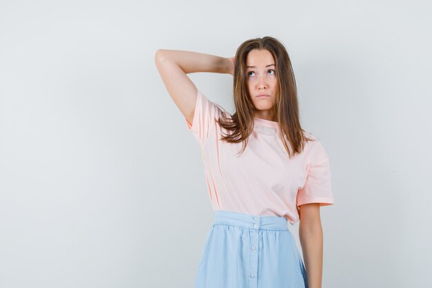 Mujer joven sosteniendo la mano detrás de la cabeza en camiseta, falda y mirando vacilante. vista frontal.