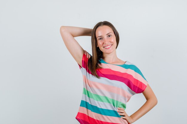 Mujer joven sosteniendo la mano detrás de la cabeza en camiseta y mirando contento, vista frontal.