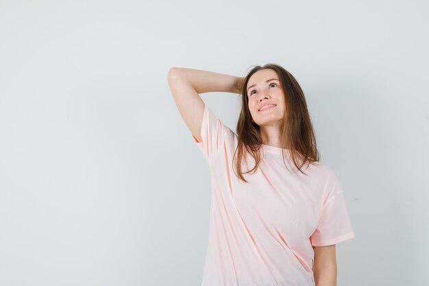 Mujer joven sosteniendo la mano detrás de la cabeza en camiseta rosa y mirando soñadora. vista frontal.