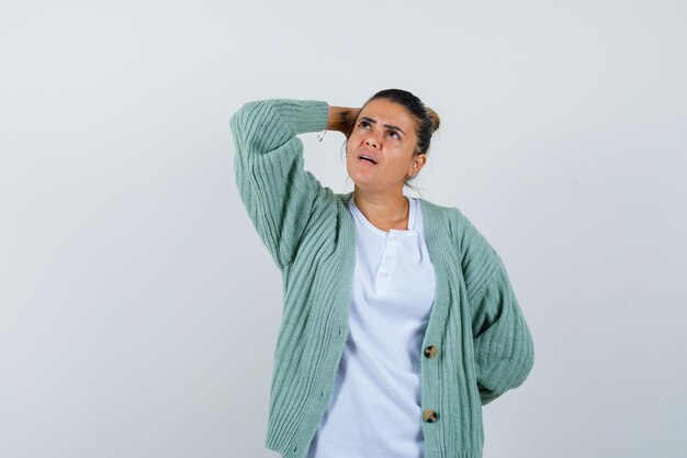 Mujer joven sosteniendo una mano detrás de la cabeza, otra mano detrás de la cintura con camisa blanca y chaqueta de punto verde menta y mirando pensativo