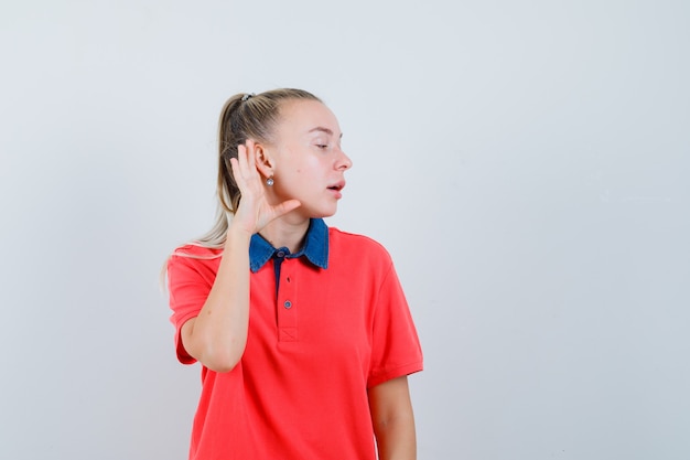 Mujer joven sosteniendo la mano detrás de la oreja en camiseta y mirando curioso