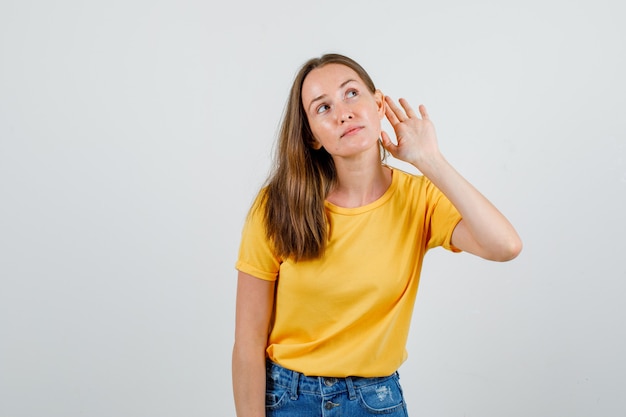 Mujer joven sosteniendo la mano detrás de la oreja para escuchar en camiseta, vista frontal de pantalones cortos.