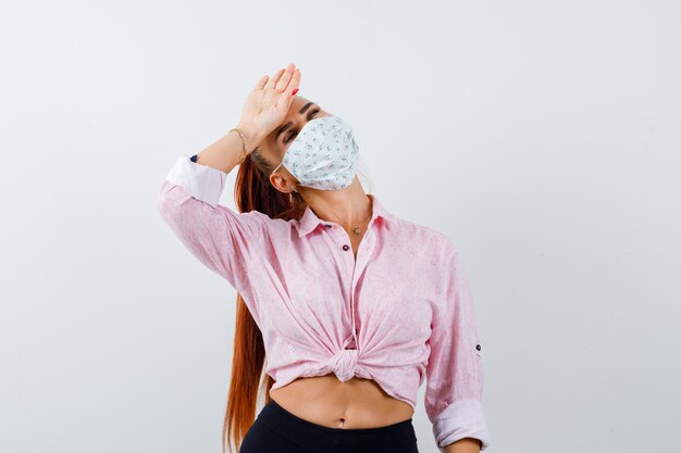 Mujer joven sosteniendo la mano en la frente en camisa, pantalón, máscara médica y mirando exhausto. vista frontal.