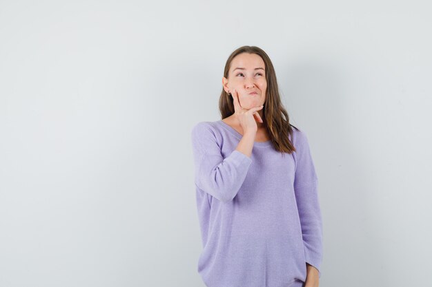Mujer joven sosteniendo la mano en la mandíbula mientras piensa en blusa lila y parece complicado