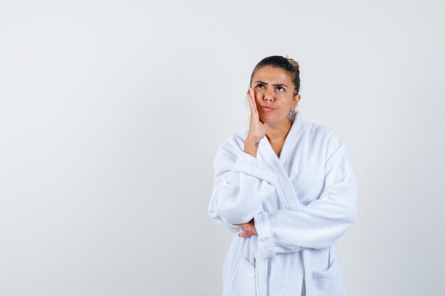 Mujer joven sosteniendo la mano en la mejilla en bata de baño y mirando pensativo