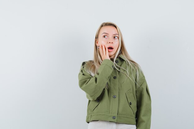 Mujer joven sosteniendo la mano en la mejilla mientras mira hacia otro lado con chaqueta verde y mira asombrada, vista frontal.