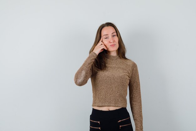 Mujer joven sosteniendo la mano en la mejilla y posando al frente en suéter dorado dorado y pantalón negro y luciendo encantador