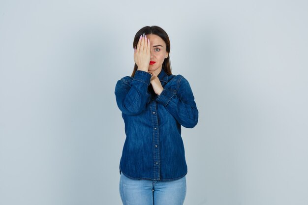 Mujer joven sosteniendo la mano en el ojo en camisa de mezclilla y jeans y mirando serio