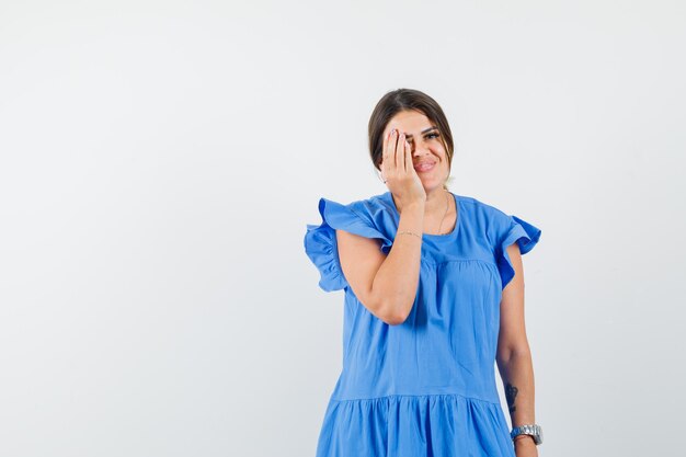 Mujer joven sosteniendo la mano en el ojo en vestido azul y mirando contento