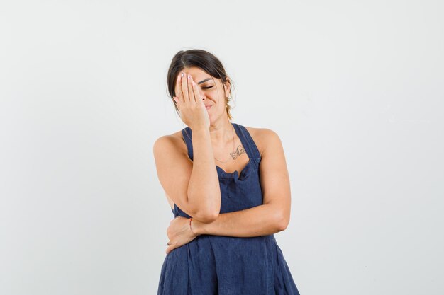 Mujer joven sosteniendo la mano en un ojo en vestido azul oscuro y con aspecto cansado