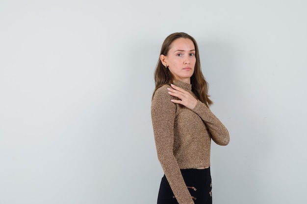 Mujer joven sosteniendo la mano en el pecho y posando al frente en suéter dorado dorado y pantalón negro y luciendo encantador