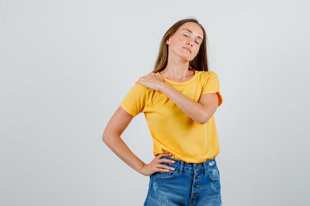 Mujer joven sosteniendo la mano sobre el hombro en camiseta, pantalones cortos y aspecto cansado. vista frontal.