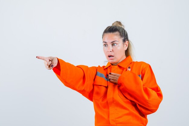 Mujer joven sosteniendo la mano sobre su pecho mientras apunta a un lado en uniforme de trabajador y mira asustada, vista frontal.