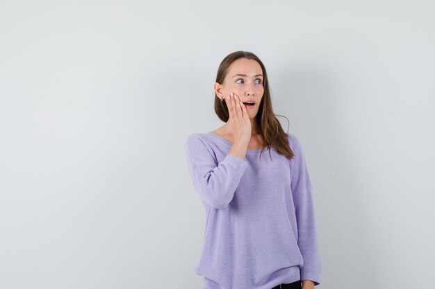 Mujer joven sosteniendo la mano sobre su rostro en blusa lila y mirando sorprendido. vista frontal. espacio para texto