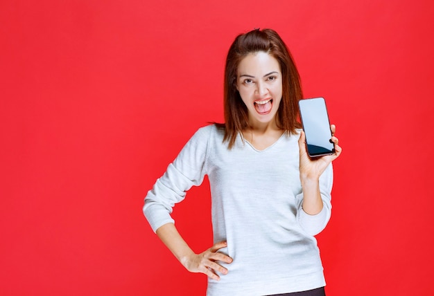 Mujer joven sosteniendo un nuevo modelo de teléfono inteligente negro y sintiéndose positivo y satisfecho