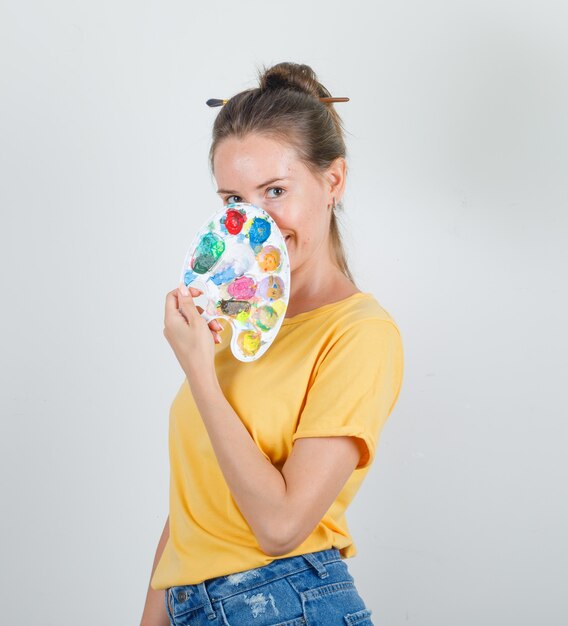 Mujer joven sosteniendo la paleta sobre la cara en camiseta amarilla, pantalones cortos de jeans y mirando alegre