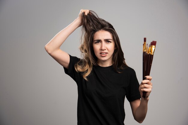 Mujer joven sosteniendo pinceles tocando su cabello en la pared gris.