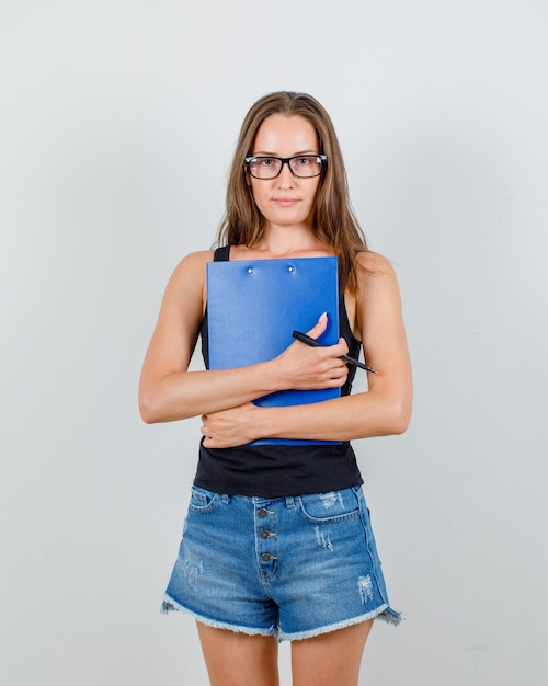 Mujer joven sosteniendo portapapeles y bolígrafo en camiseta, pantalones cortos, gafas y mirando confiado. vista frontal.