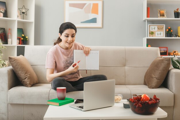 Mujer joven sosteniendo portátil sentado en el sofá detrás de la mesa de café mirando portátil en la sala de estar