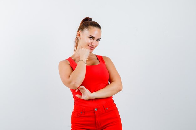 Mujer joven sosteniendo el puño en la barbilla en camiseta sin mangas roja, pantalones y aspecto atractivo, vista frontal.