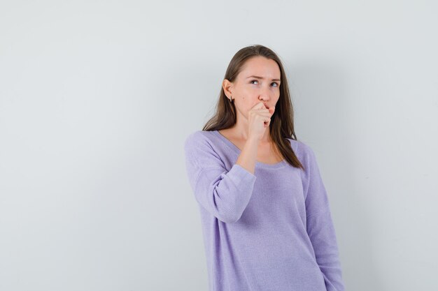 Mujer joven sosteniendo el puño en la boca mientras mira hacia otro lado en blusa lila y mira pensativa
