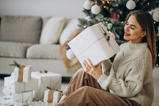 Mujer joven sosteniendo regalos de Navidad y sentado bajo el árbol de Navidad