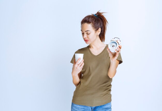 Mujer joven sosteniendo un reloj despertador y una taza de bebida apuntando a la rutina matutina