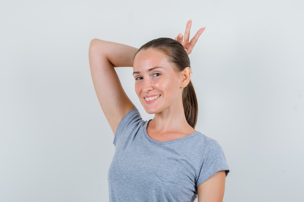 Foto gratuita mujer joven sosteniendo el signo v detrás de la cabeza como cuernos en camiseta gris y mirando divertido, vista frontal.