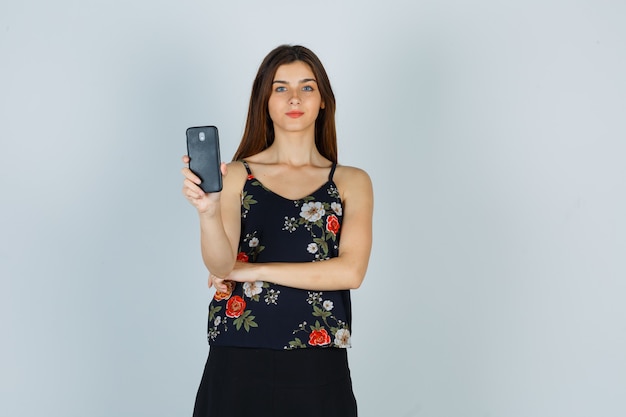 Mujer joven sosteniendo smartphone en blusa, falda y mirando complacido. vista frontal.