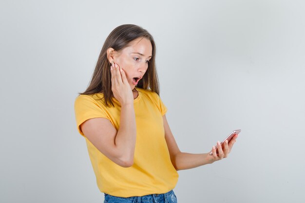 Mujer joven sosteniendo smartphone en camiseta amarilla, pantalones cortos y mirando sorprendido