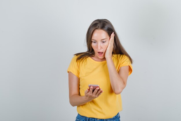 Mujer joven sosteniendo smartphone en camiseta amarilla, pantalones cortos y mirando sorprendido