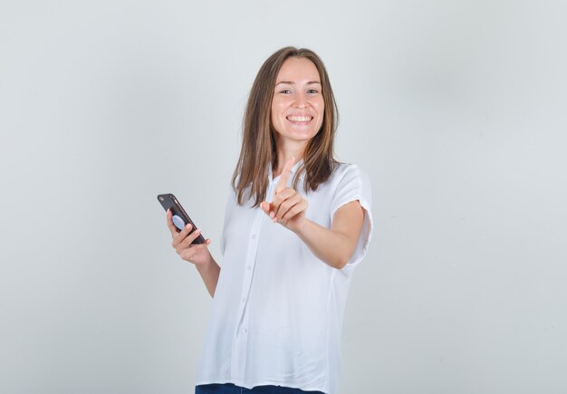 Foto gratuita mujer joven sosteniendo smartphone y gesticulando no con el dedo en camisa blanca