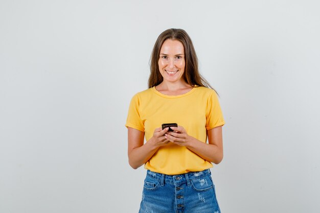 Mujer joven sosteniendo smartphone y sonriendo en camiseta, vista frontal de pantalones cortos.