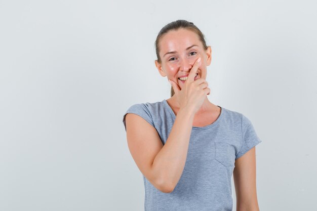 Mujer joven sosteniendo su barbilla en camiseta gris y mirando alegre, vista frontal.