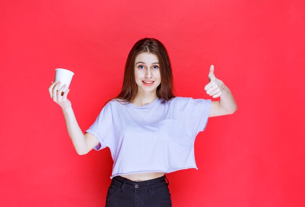 mujer joven sosteniendo una taza de agua desechable blanca y mostrando el signo de satisfacción.