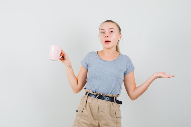 Mujer joven sosteniendo una taza de bebida en camiseta, pantalón y mirando confundido, vista frontal.