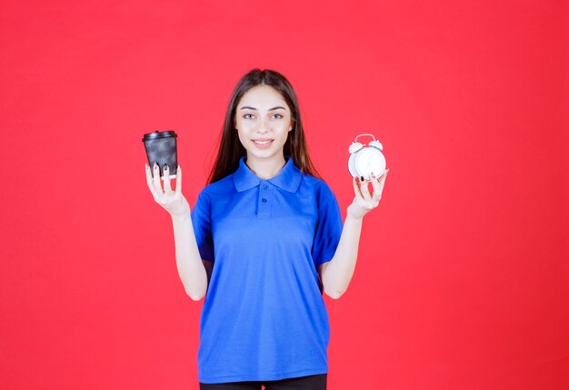 Mujer joven sosteniendo una taza de bebida desechable negra y un reloj despertador