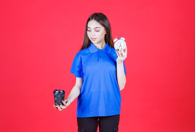 Mujer joven sosteniendo una taza de bebida desechable negra y un reloj despertador