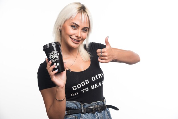 Mujer joven sosteniendo una taza de café en la pared blanca mostrando los pulgares para arriba.