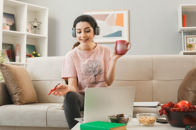 Mujer joven sosteniendo una taza de té con lápiz usando audífonos portátil usado sentado en el sofá detrás de la mesa de café en la sala de estar