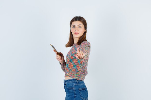 Mujer joven sosteniendo el teléfono, apuntando al frente en géneros de punto, jeans y mirando serio, vista frontal.