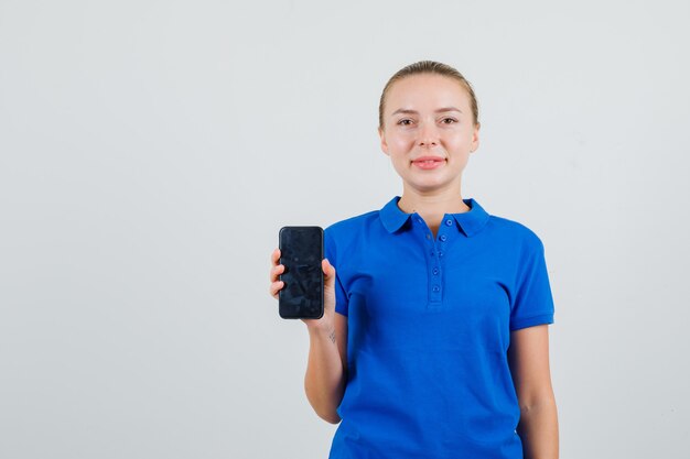 Mujer joven sosteniendo teléfono móvil en camiseta azul y mirando complacido