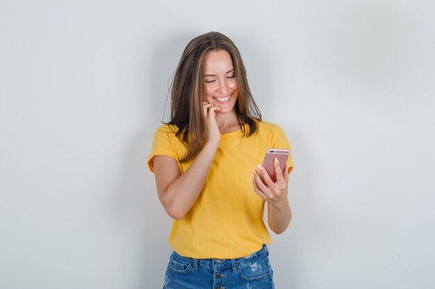 Mujer joven sosteniendo el teléfono móvil con los dedos en la cara en camiseta, pantalones cortos y mirando feliz