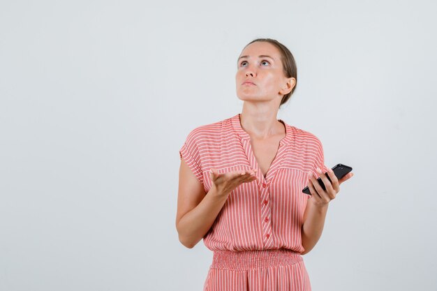 Mujer joven sosteniendo el teléfono móvil mientras mira hacia arriba con un vestido a rayas y parece confundido, vista frontal.