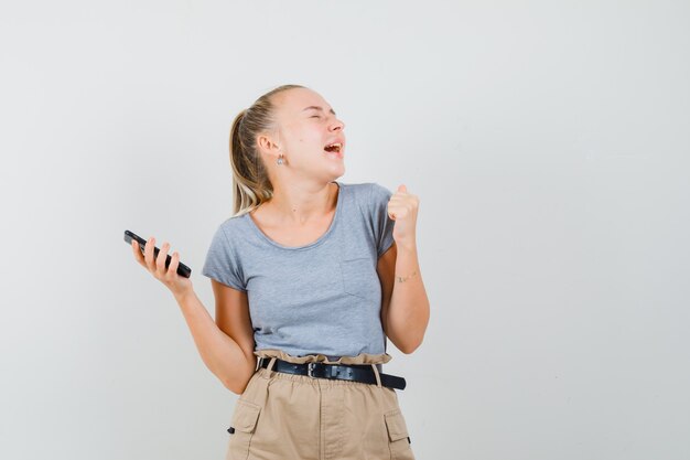 Mujer joven sosteniendo el teléfono móvil, mostrando el gesto de ganador en camiseta, pantalón y luciendo dichoso. vista frontal.