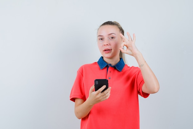 Mujer joven sosteniendo el teléfono móvil, mostrando gesto ok en camiseta y mirando confiado