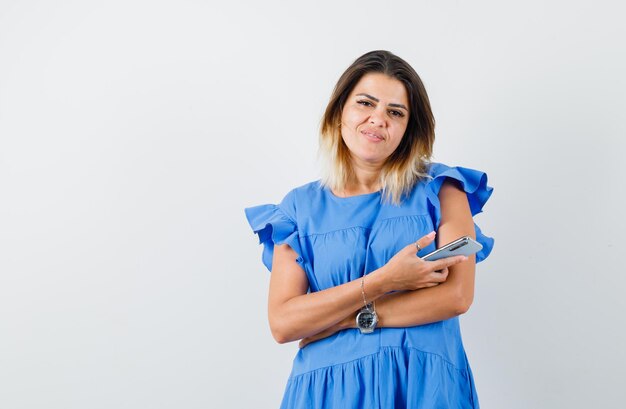 Mujer joven sosteniendo teléfono móvil en vestido azul y mirando complacido