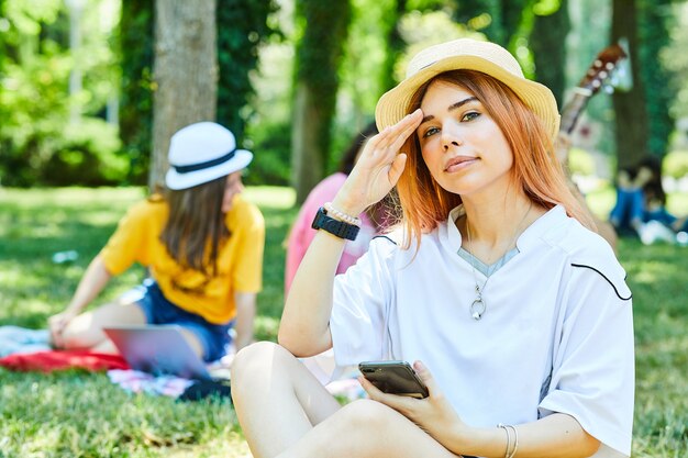 Una mujer joven sosteniendo un teléfono y sentada en el césped con sus amigos en la espalda.