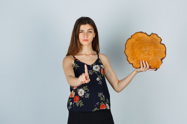 Mujer joven sosteniendo un trozo de madera, mostrando un gesto de espera en la blusa, falda y mirando serio, vista frontal.