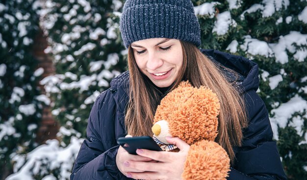 Una mujer joven sostiene un oso de peluche y un teléfono inteligente en sus manos en tiempo de nieve